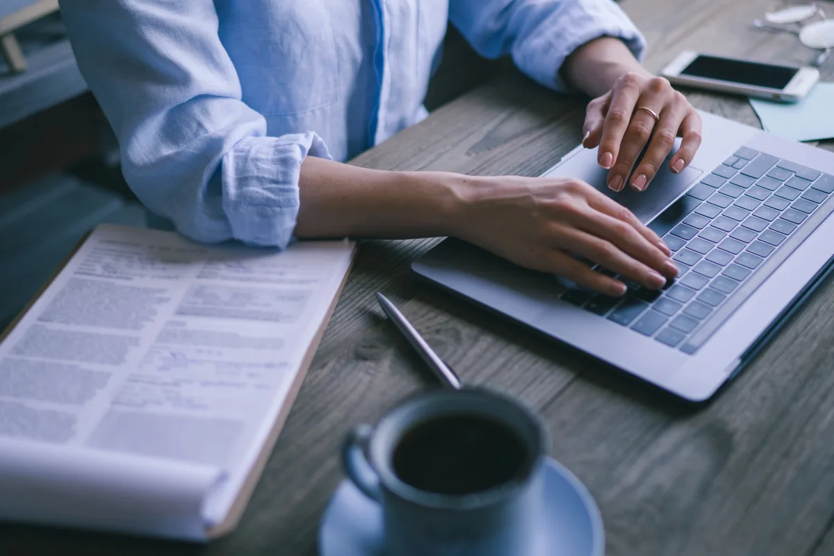 Close-up of a writer typing on a laptop at a wooden table, with a printed document, a pen, a cup of black coffee, and a smartphone nearby