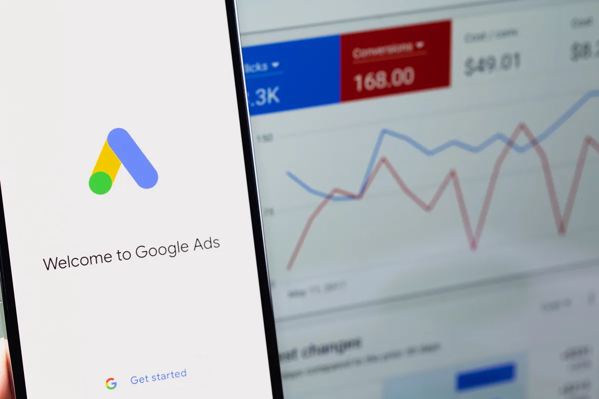 Smartphone showing the Welcome to Google Ads screen in front of a monitor displaying a Google Ads dashboard with clicks, conversions, and a performance chart
