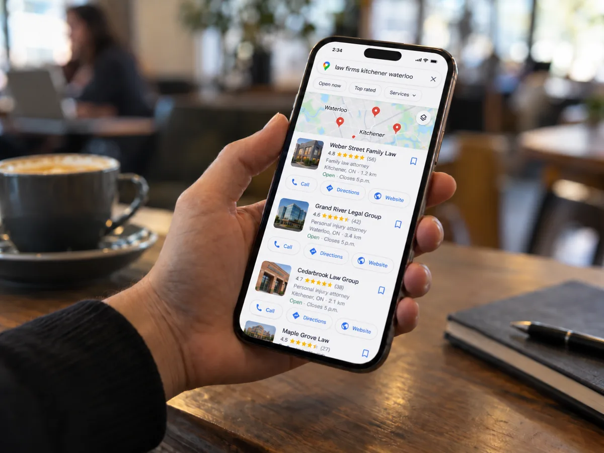 Hand holding a smartphone in a café, showing a Google Maps-style results page for fictional law firms in Kitchener-Waterloo, with a coffee cup and notebook blurred in the foreground.