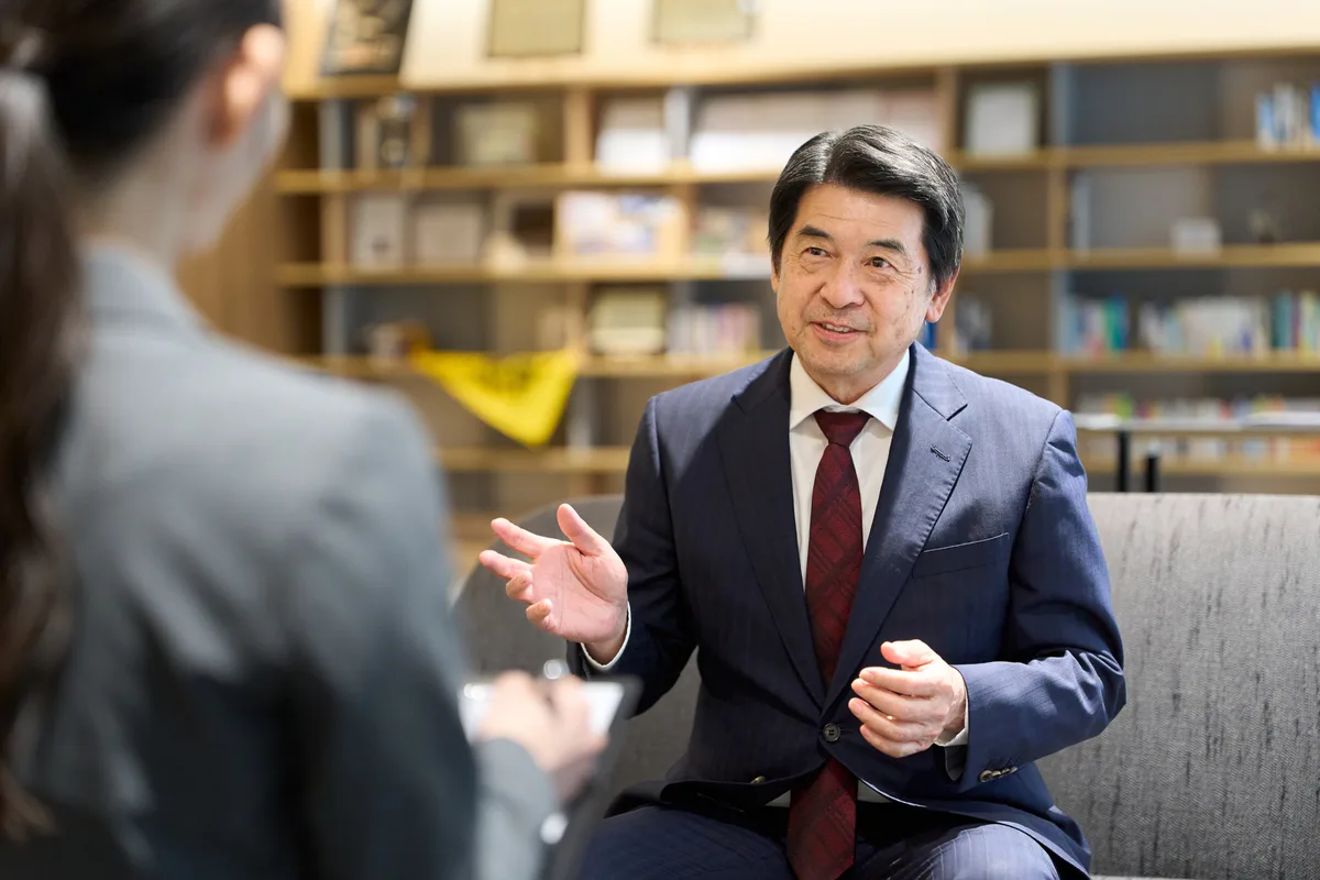 Lawyer in a navy suit and red tie gesturing mid-answer to a reporter taking notes on a tablet, seated in an office lounge in front of bookshelves