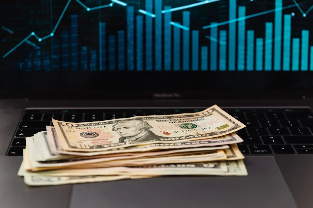 A stack of USA currency on a laptop keyboard, with the screen displaying blue line graphs and bar charts.