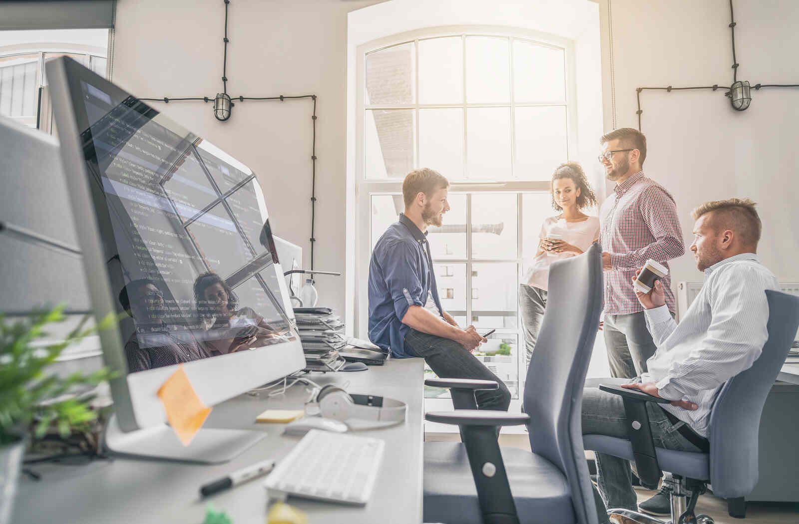 A website design and development team gathers for a meeting near a computer showing lines of HTML code.