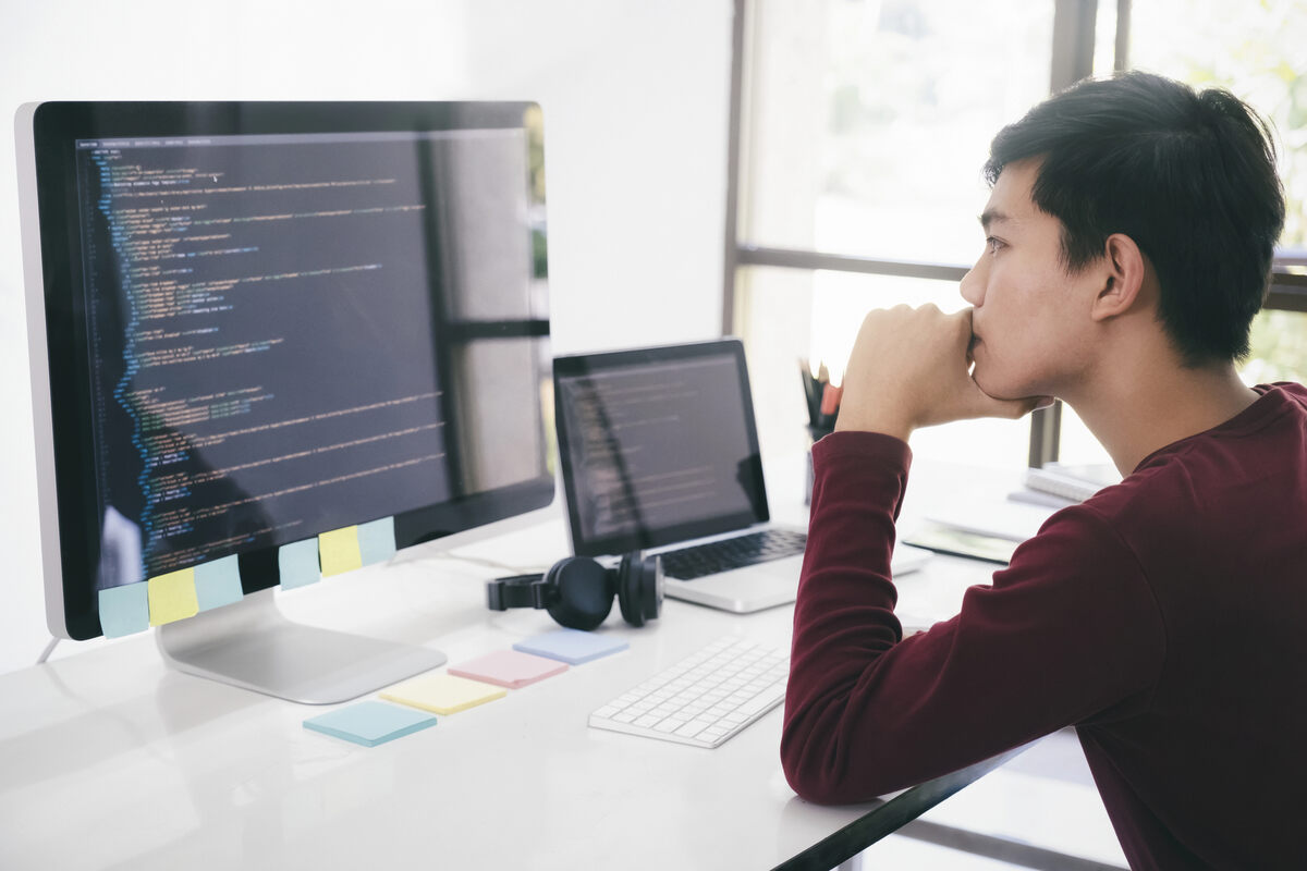 A software engineer analyzes HTML code on his dual monitor setup in a clean and bright office.