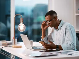 A young professional contemplating ideas as he uses his laptop and mobile phone in an office during the evening.