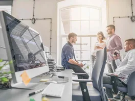 A website design and development team gathers for a meeting near a computer showing lines of HTML code.
